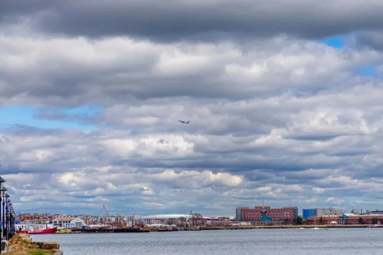 View of Boston Harbor with a cloudy sky, cityscape, and a distant airplane.