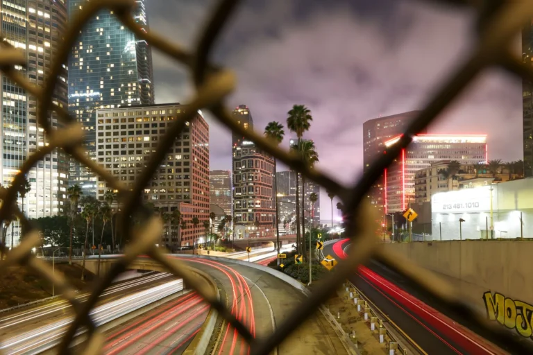 Long exposure night shot capturing the dynamic Los Angeles cityscape through a chain-link fence.