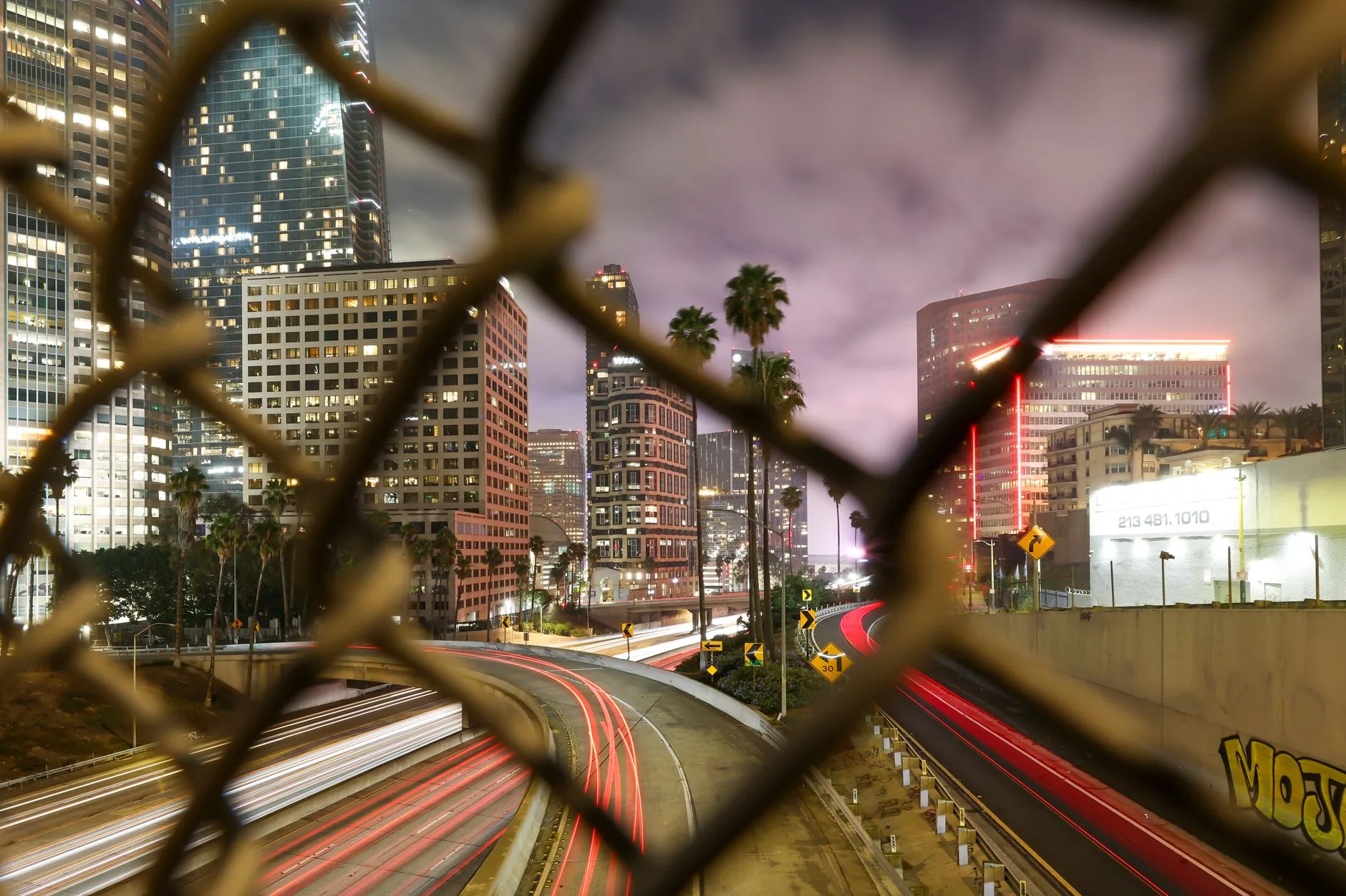 Long exposure night shot capturing the dynamic Los Angeles cityscape through a chain-link fence.