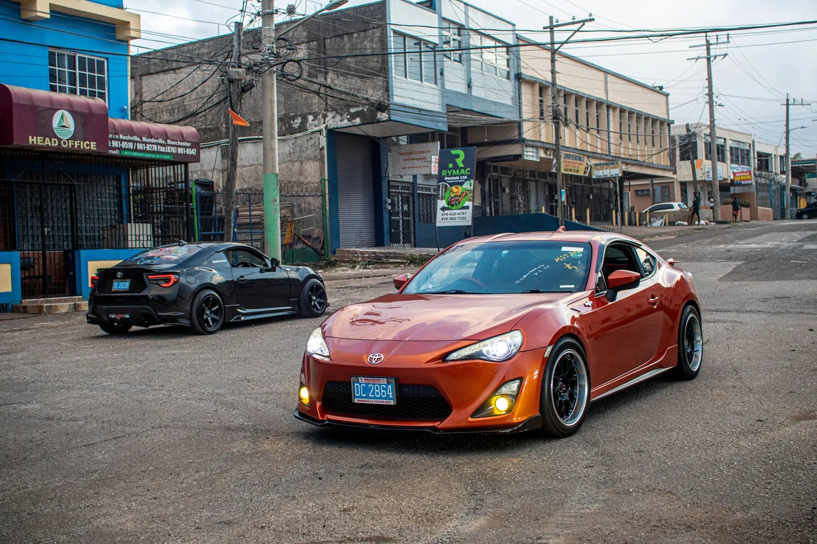 Two modern sports cars parked on a rustic street in Jamaica, showcasing contrast between sleek design and urban setting.