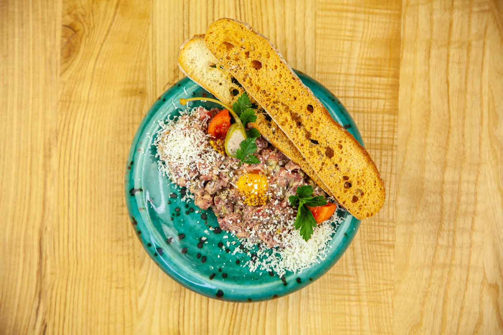 Delicious beef tartare plated with cheese, tomato, and crispy bread on a turquoise dish.