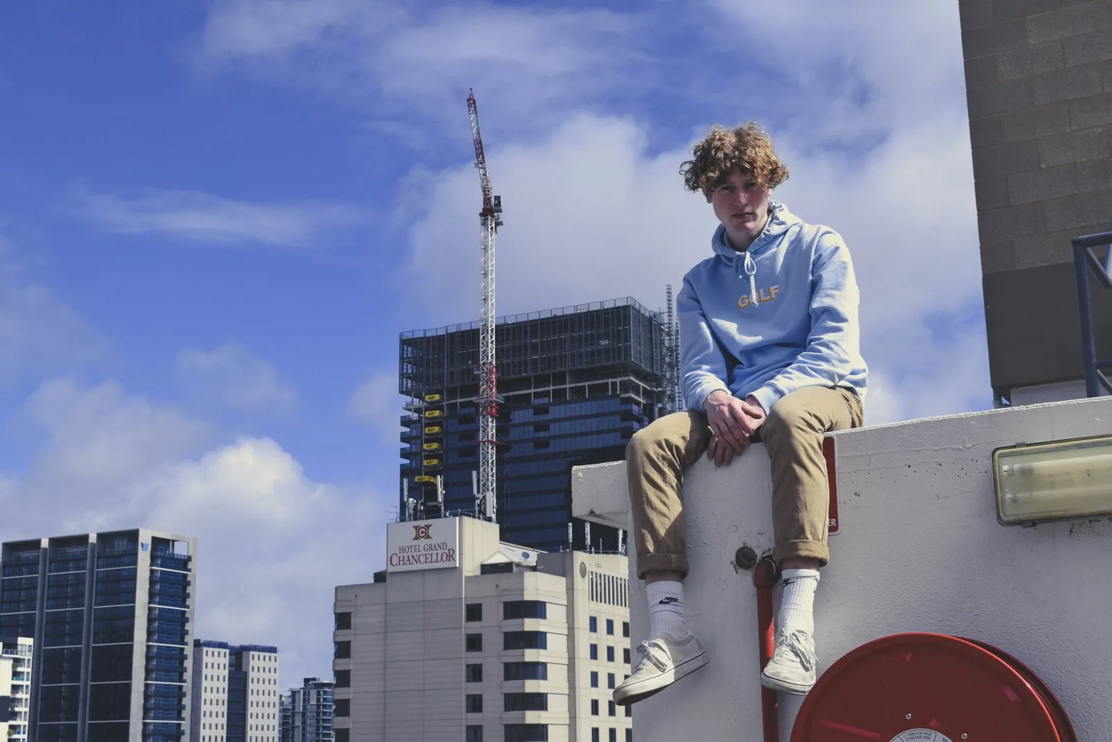 Young man in blue hoodie sitting against Adelaide city skyline under a clear sky.