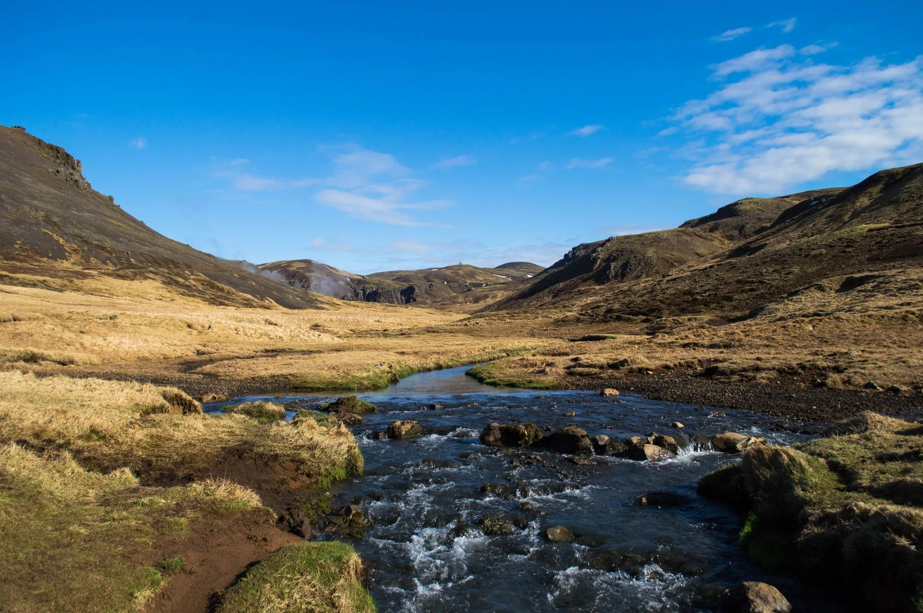 A picturesque view of a river flowing through a tranquil Icelandic valley surrounded by hills under a clear blue sky.