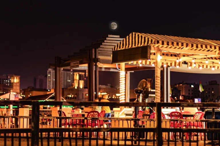 Nighttime view of a lively outdoor cafe in Dubai with a full moon and city skyline in the background.